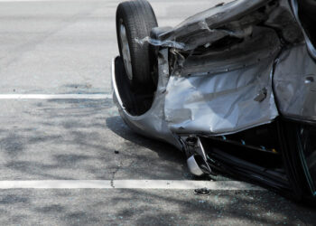 A silver car just involved in a car crash at an intersection of a city street, upside down and against the curb.