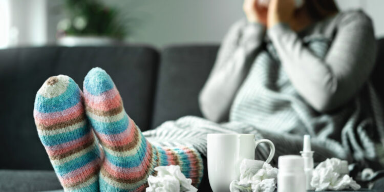 Sick woman with flu, cold, fever and cough sitting on couch at home. Ill person blowing nose and sneezing with tissue and handkerchief. Woolen socks and medicine. Infection in winter. Resting on sofa.
