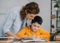 beautiful woman standing near adorable writing in copy book while doing schoolwork at home